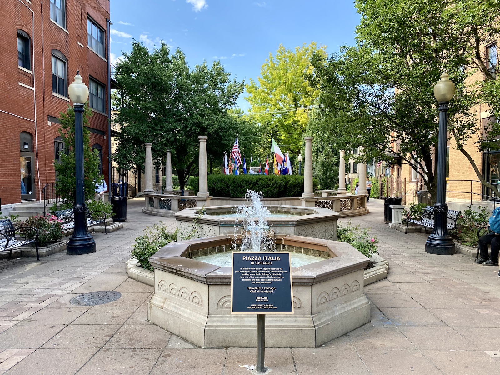 A plaque stands in front of the fountains at Piazza Italia Di Chicago, flags in the distance surround the fountains in the distance.