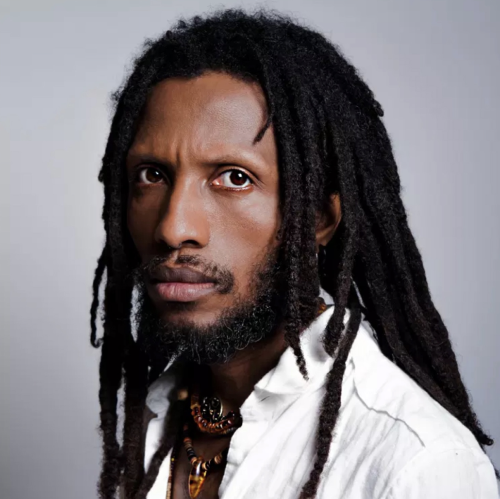 Portrait of Kao Ra Zen. A black man with long black dreads, short beard and mustache, gazing with deep brown eyes into the camera, Zen is wearing a white collared shirt and a beaded colorful necklace. 